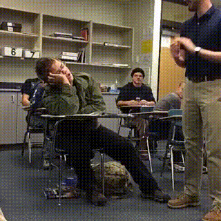 Video gif. A candid video of a student who has fallen asleep in class. The teacher comes by and starts clapping to prank him and he startles awake, mindlessly clapping along with the teacher as if he was never asleep.