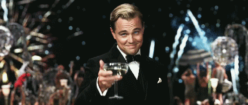 Movie gif. Wearing a tuxedo, Leonardo DiCaprio toasts to us with a glass of white wine and a complicated smile. A large celebration with fireworks and a ferris wheel goes on behind him.