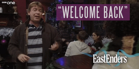 TV gif. Adam Woodyatt as Ian Beale from Eastenders stands in a room adorned with Christmas decor and throws his hands up in mock excitement to emphasize his sarcasm while he says, "Welcome back," which appears as text.