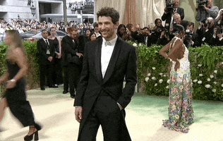 Met Gala 2024 gif. Josh O'Connor wearing a Loewe tuxedo sans tie, poses childishly, smiling happily for the cameras.