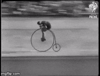Video gif. An old black and white video of a bicycle race. Men pedal fast along a curved race track on old timey bicycles that have one big wheel in the front.