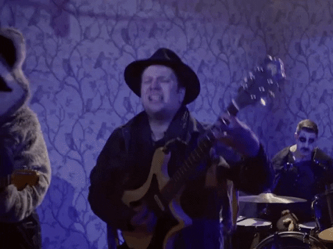 Video gif. Patrick Stump from Fall Out Boy plays his guitar on stage, singing energetically into the mic against a purple wallpaper background. 