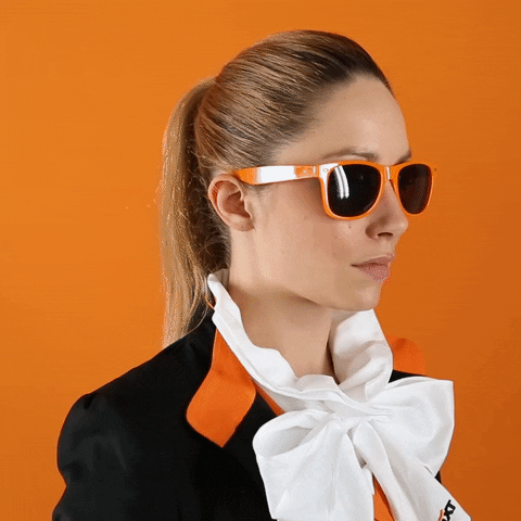 Video gif. Woman wearing a black, orange, and white outfit against an orange background turns to look at us, slowly lowers her orange sunglasses, and looks at us seriously.