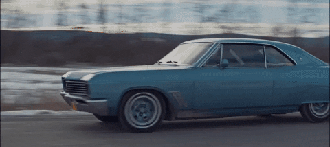 Music Video gif. Blue old car in Tei Shi’s How Far music video drives backwards and then spins around on a road.