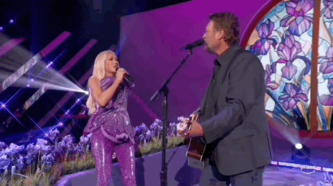 Academy of Country Music Awards gif. Gwen Stefani and Blake Shelton perform as a couple. The back of Shelton's faces the camera as strums the guitar strapped around his body and lovely looks at Stefani who maintains eye contact as she sings into a handheld mic with Blake. Gwen, wearing a sparkly purple outfit that matches the stage make a "C" shape with her hand and closes it like a crab claw in front of Shelton's face in a teasing manner.