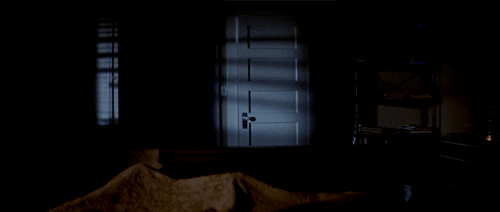 Movie gif. In a scene from Halloween, a person lies in a dark room as the door slowly opens, revealing a tall figure dressed as a ghost, draped in a white sheet and wearing sunglasses.