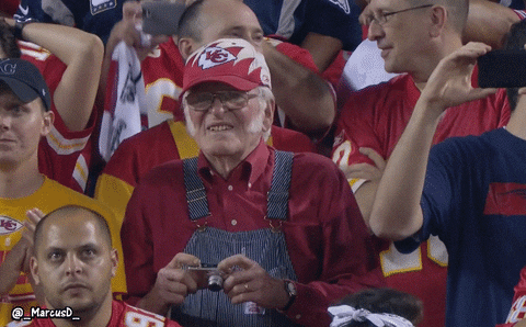 Sports gif. Older man stands pridefully in the football stands, wearing striped overalls with a red collared shirt and a Kansas City Chiefs cap. He slowly brings up his digital camera to capture the exciting moment. Text appears at the bottom and reads, "Neat!"
