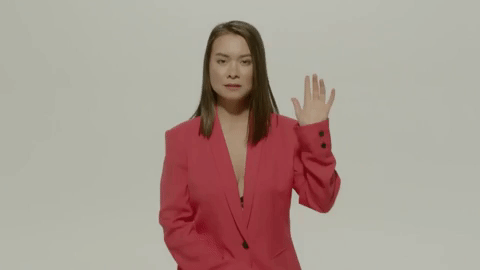 Celebrity gif. Mitski is sitting on a stool with a blank expression and she has one hand up to give a slight wave.