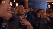 TV gif. Fans in the audience of Titan Games are all standing up and cheering raucously, clapping their hands and waving their arms in the air.