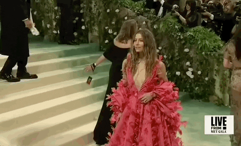 Met Gala 2024 gif. We zoom in on Jessica Biel waving and smiling at camera, wearing a hot pink Tamara Ralph Couture full-length gown with a deep-v neckline. The full billowing skirt flares out to a dramatic train. She wears a billowing shrug that matches the pattern of her dress around her back and arms.  