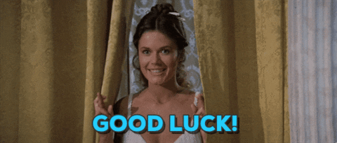 Video gif. Brunette woman stands between yellow drapes and grins, holding up both hands with fingers crossed. Text, "good luck!"