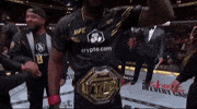 Sports gif. Leon Edwards, a UFC fighter, walks across the ring, making a bicep and pointing at us. Joe Rogan is standing next to him with a microphone as others fill the ring behind him. 