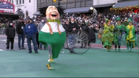 Video gif. Two armless munchkins in costume from Wicked jot through the Macy's Thanksgiving parade.