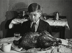 Video gif. A black and white video of a young boy licking his lips as he rocks back and forth in anticipation behind a thanksgiving turkey on a dining table. 