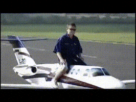 Video gif. Boy sits on a mini airplane that rolls around the tarmac slowly.
