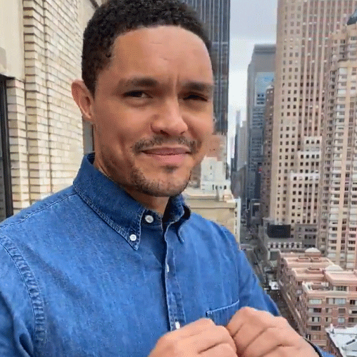 Celebrity gif. On a balcony with skyscrapers in the background, Trevor Noah smiles earnestly and holds his hands to his chest in a heart shape