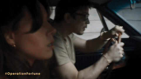 Movie gif. Swerving in a red Mustang, Aubrey Plaza as Sarah and Josh Hartnett as Danny in Operation Fortune: Ruse de Guerre frantically try to outrun a group of men shooting rifles from a black jeep.