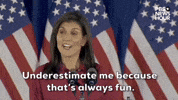 Political gif. Nikki Haley shrugs and shakes her head with a smile as she stands in front of a curtain of American flags, saying, "Underestimate me because that's always fun."