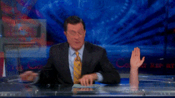 Late Show gif. Hand pops up from under Stephen Colbert's desk and he slaps it, high fiving them before turning back to grin at us with satisfaction.