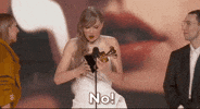 Celebrity gif. Taylor Swift holds a Grammy on the awards stage as she smiles and covers her mouth in surprise, turning away from the microphone. Text, "No!"