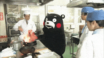 Video gif. From a distance, two chefs observe another chef stirring food on fire in a wok, while the Kumamon mascot waves its hand in fright and trembles.