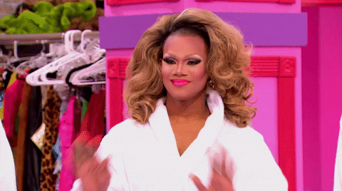Reality TV gif. Chi Chi Devayne on Rupaul’s Drag Race wears a white robe. She waves with both hands, and smiles. 