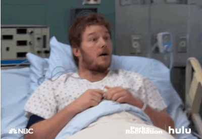 Parks and Recreation gif. Chris Pratt as Andy lays in a hospital bed as a patient and looks out expectantly, giving a thumbs up.