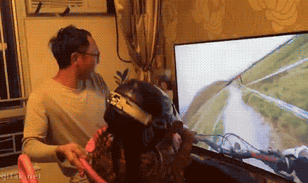 Video gif. Father lifts his daughter in front of the TV as she rides on her toy pink motorcycle while wearing a big motorcycle helmet. The TV shows a first person perspective video of someone riding their bike on a dirt path. The father jerks her left and right to simulate the movements in the video.