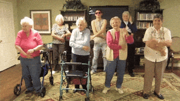 Video gif. A bunch of senior women stand in a living room, along with one young man in sunglasses and a tank top. A few of the older women have walkers, and they all stand together doing the macarena.