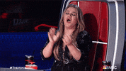 TV gif. Kelly Clarkson on The Voice. She's sitting in her judges chair and she claps enthusiastically while whooping.