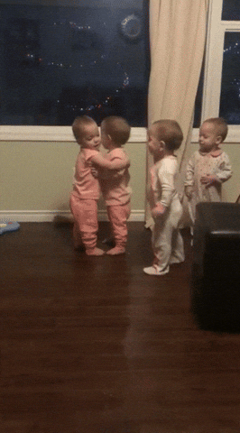 Video gif Four babies all stand up Two babies in pink hug each other while the other two awkwardly stand and wait their turn One of the babies in white taps a baby in pink and they hug The four babies then go around hugging each other and smiling with excitement as they pat each others backs