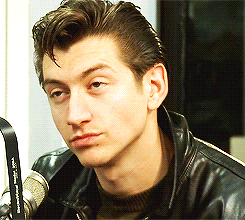 Video gif. Man in a leather jacket with gelled hair sits in front of a microphone while chewing on something slowly, appearing completely indifferent.