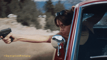 Movie gif. Aubrey Plaza as Sarah in Operation Fortune: Ruse de Guerre. She's riding in the passenger seat of a drifting car and she shoots a gun out the window, with a determined expression on her face.