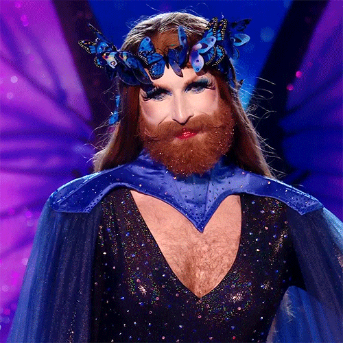 Reality TV gif. Drag Queen Gingzilla on the show Queen of the Universe with long lashes and a long beard wears a crown made up of blue butterflies, a blue cape, and a sparkly dress. They place their well manicured hand over their chest in appreciation and smile, nodding and batting away tears from their eyes.