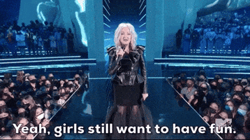 Celebrity gif. Cyndi Lauper performing at the 2021 VMAs. She is on stage and holds a microphone in her hand as she announces, "Yeah, girls still want to have fun," and the crowd around her cheers when she points at them.
