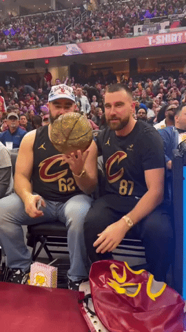 Sports gif. Travis and Jason Kelce sit in the audience at a Cleveland Cavaliers game wearing Cavaliers shirts. Jason tosses a bedazzled Cavaliers basketball in one hand and Travis throws up a shaka. Both pose for the camera.