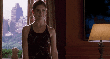 Movie gif. Sarah Michelle Gellar as Kathryn in Cruel Intentions. She's standing by a window and she puts her hand up to her face to give a little wave at someone who walks in.