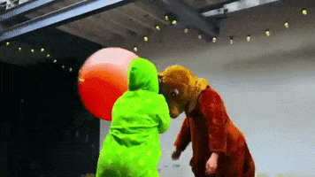 Video gif. Girl in a onesie swings a red hooper ball and hits a person in a kangaroo costume. The Kangaroo stumbles back from the blow.