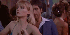 Movie gif. Michelle Pfeiffer as Elvira Hancock in Scarface Rumbas with stoic sex appeal, Al Pacino as Tony Montana bopping up behind her with awkward machismo.