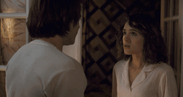 Stranger Things gif. Natalia Dyer as Nancy Wheeler and Charlie Heaton as Jonathan Byers stare at each other intensely in a dimly lit room before passionately kissing and holding each other in a tight embrace.