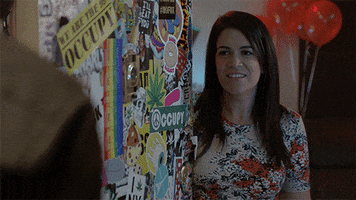 TV gif. Abbi Jacobson as Abbi on Broad City leans on a sticker filled wall. She nods her head up coolly, saying “How you doin’,” in an awful attempt at flirting.