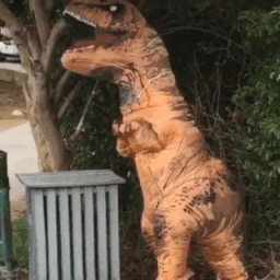 Video gif. A person wearing an inflatable T-Rex costume is standing in front of a bush and eagerly waves with their short, stubby paw. 