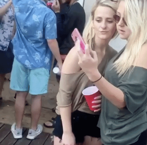 Video gif. Two women are at a party and about to take a selfie when one woman sees an uncomfortable text her friend has just received. She pulls a face and looks at us with a slanted lip.