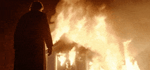 Movie gif. Brendan Gleeson as Father James in Calvary stands in the foreground, watching a small building engulfed in flames.