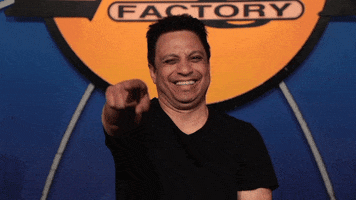 Video gif. Comedian Johnny Sanchez sits in front of a Laugh Factory sign. He’s laughing hard with his eyes closed, leaning back and pointing at us.