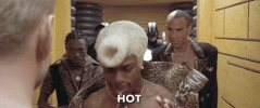 Movie gif. Followed by his entourage, Chris Tucker as Ruby in The Fifth Element struts towards us with a wild blond hairdo, wearing leopard print, and yells into his headset microphone, “HOTTT!”
