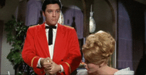 Celebrity gif. Elvis Presley nonchalantly pops a bottle, but champagne violently explodes out and hits a woman in the face.