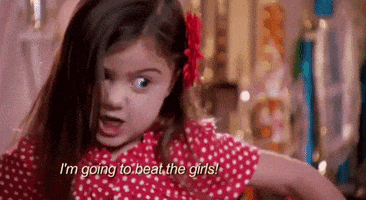Reality TV gif. In a clip from Toddlers in Tiaras, a little girl wearing a red polka-dot dress and a flower in her hair makes an intensely angry side-eye at someone offscreen. Text, "I'm going to beat the girls!"