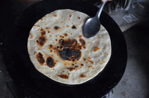 A round piece of bread (roti) is being cooked poorly on a flat plan on the stove.