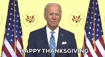 Political gif. Joe Biden stands in front of a yellow wall flanked by an American flag on each side and says, “Happy Thanksgiving.”